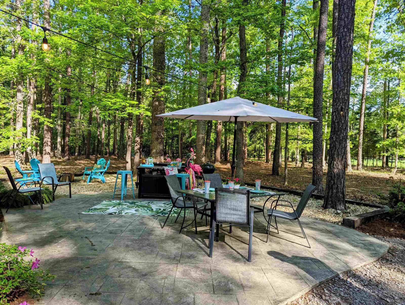 A tranquil outdoor patio setting with a dining table, chairs, and an umbrella surrounded by a lush forest.