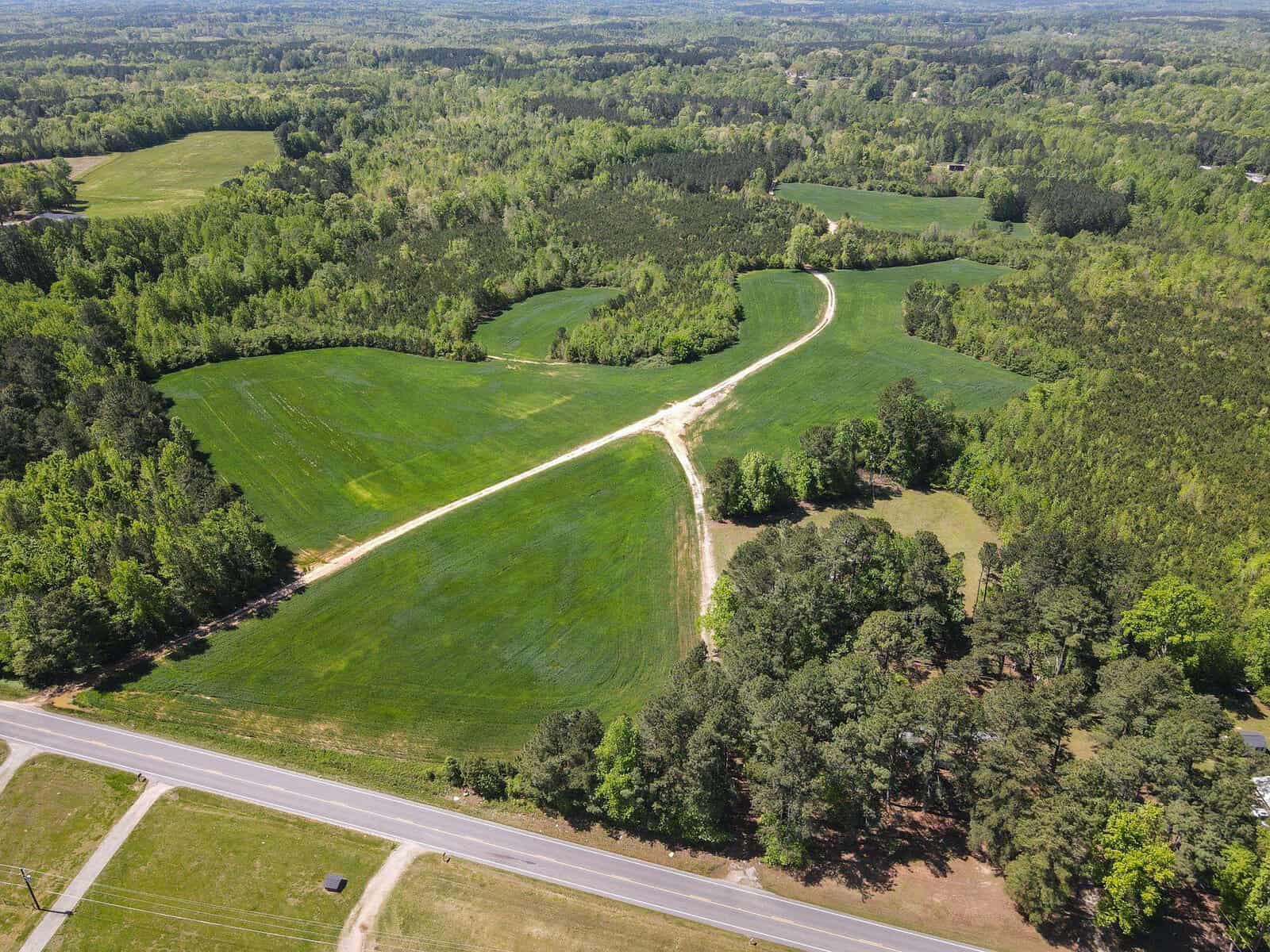 Aerial view of a rural landscape showing a green field with a dividing path, surrounded by trees and a road to the bottom of the image.