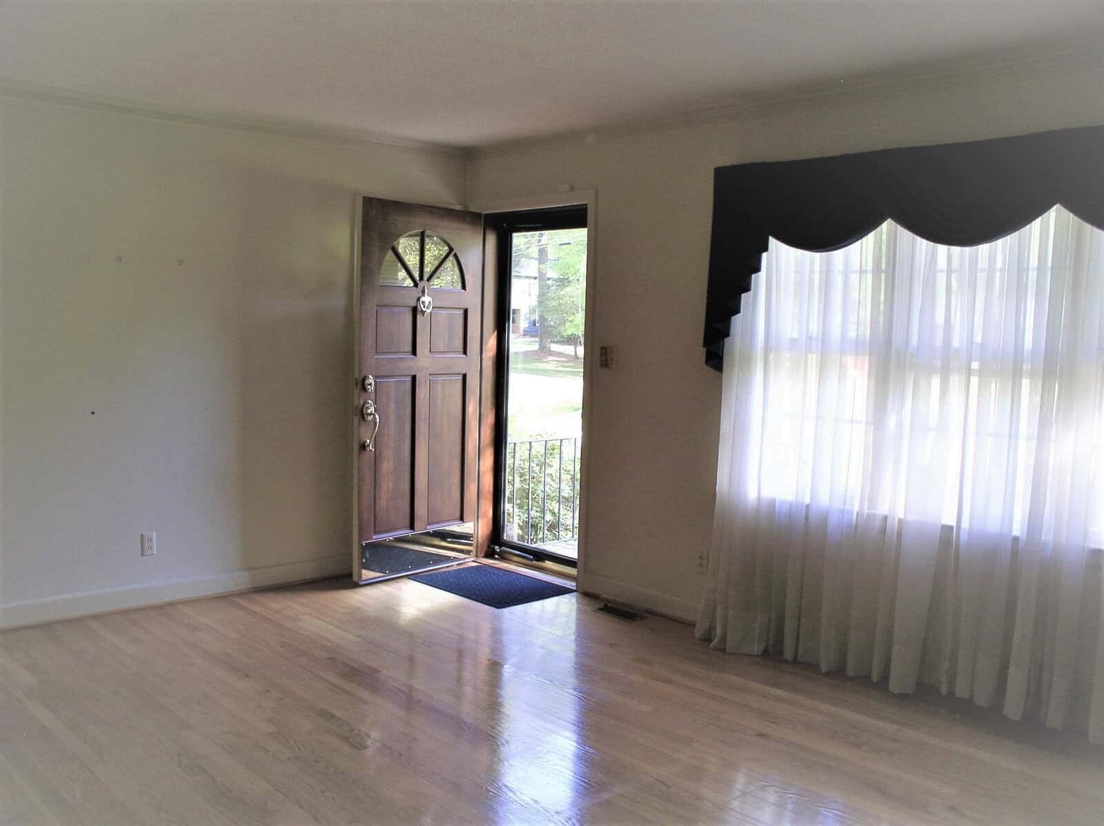 An empty room with laminate flooring, a half-opened front door with glass panels, and a large window draped with sheer curtains, allowing natural light to enter.