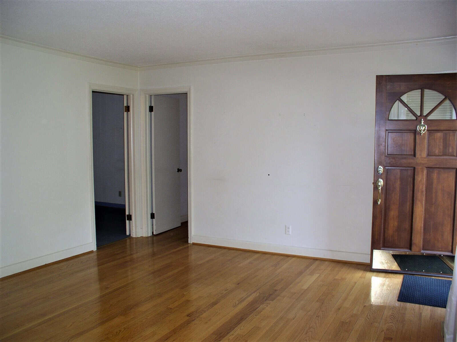 An empty room with wooden flooring, featuring a closed brown front door with a curved window design at the top, and an open interior door leading to another room.