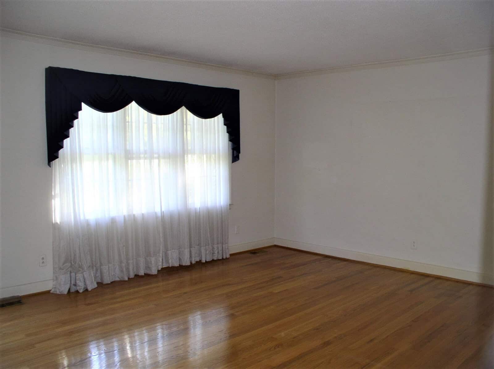 A spacious empty room with hardwood flooring, white walls, and a large window covered by sheer curtains and a navy blue valance.