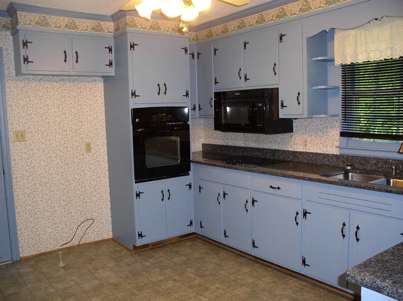 A dated kitchen interior with blue cabinets, speckled countertops, and white appliances. wallpaper borders adorn the top of the walls.