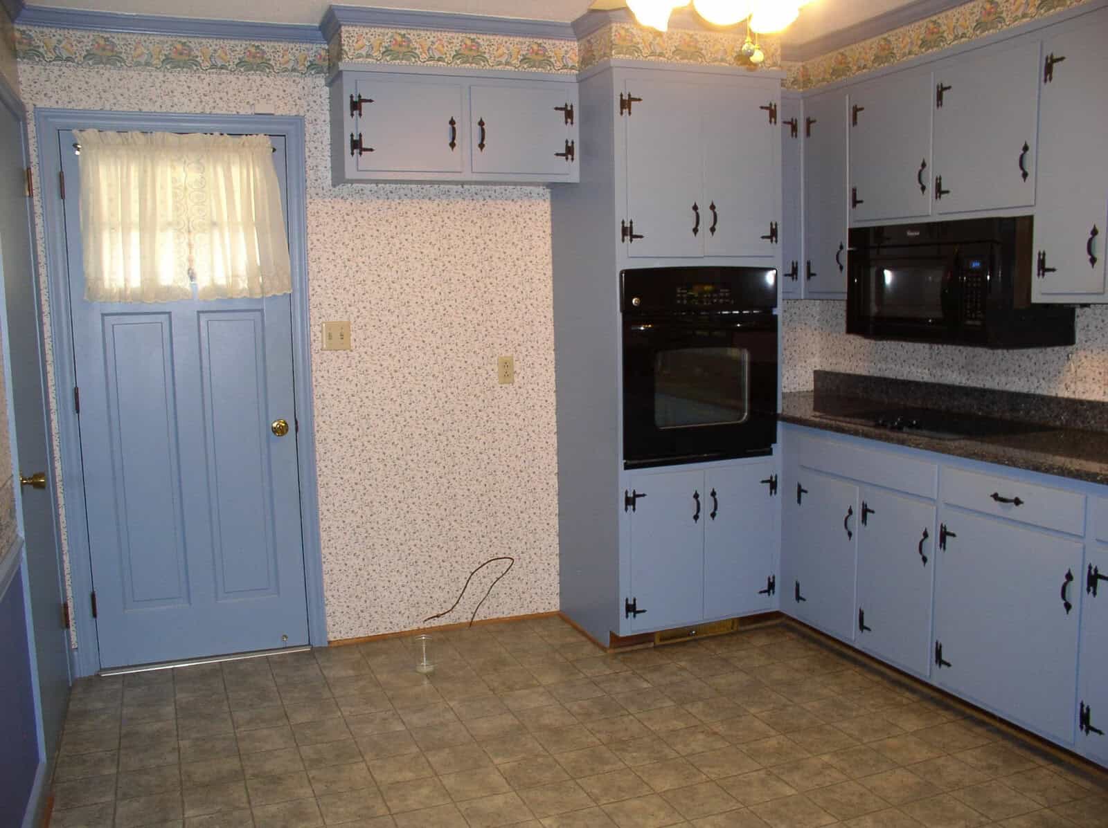 A traditional kitchen with blue cabinetry, white appliances, floral wallpaper border, and a linoleum floor.