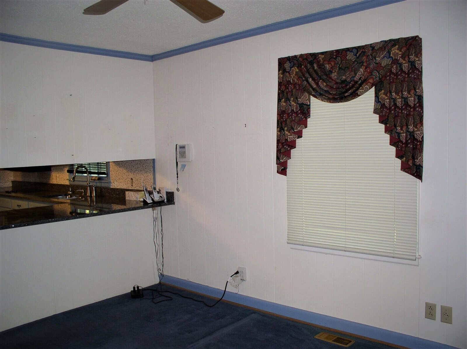 Interior of a room with white walls, a window with a patterned valance, and a view into a kitchen with brown countertops. there is a thermostat on the wall and an electrical cord on the floor.