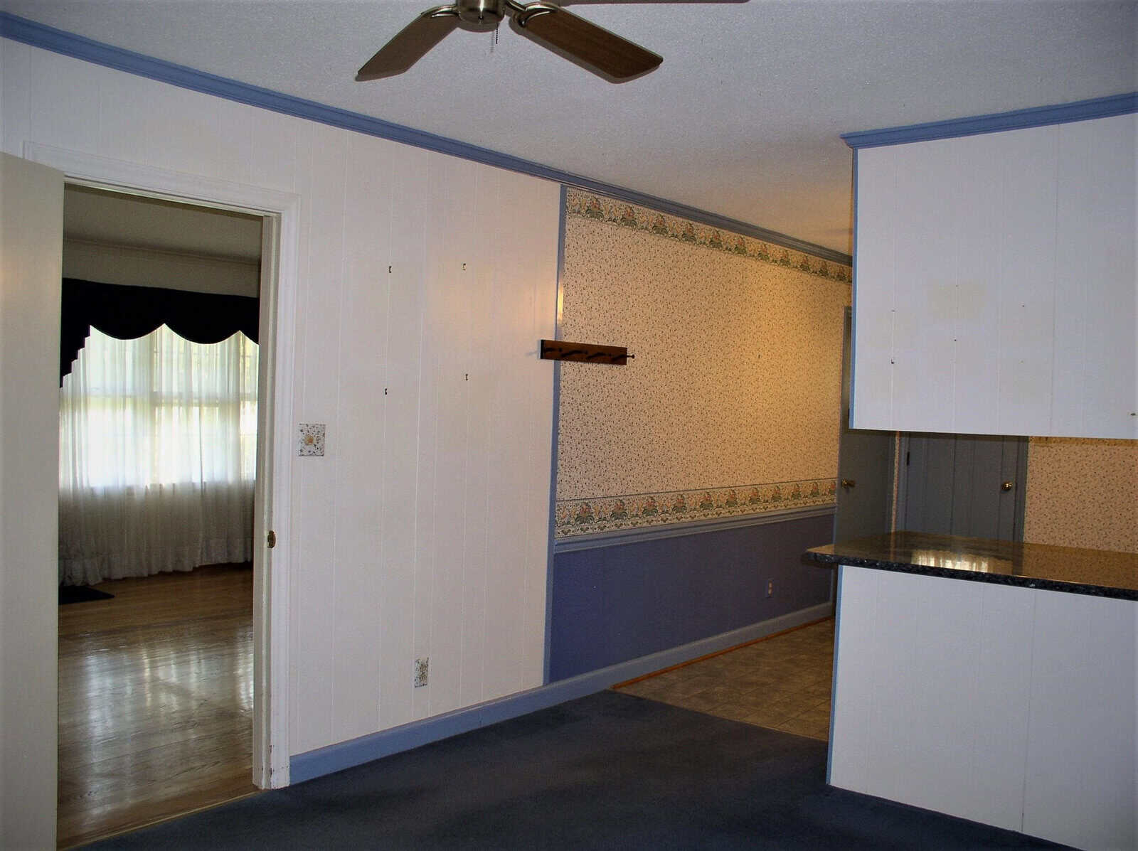 An empty room featuring a kitchen area with white cabinets and a countertop on the right, and an adjacent living space with a curtained window and a ceiling fan. the wall has a wallpaper border and the floors are partially carpeted and tiled.