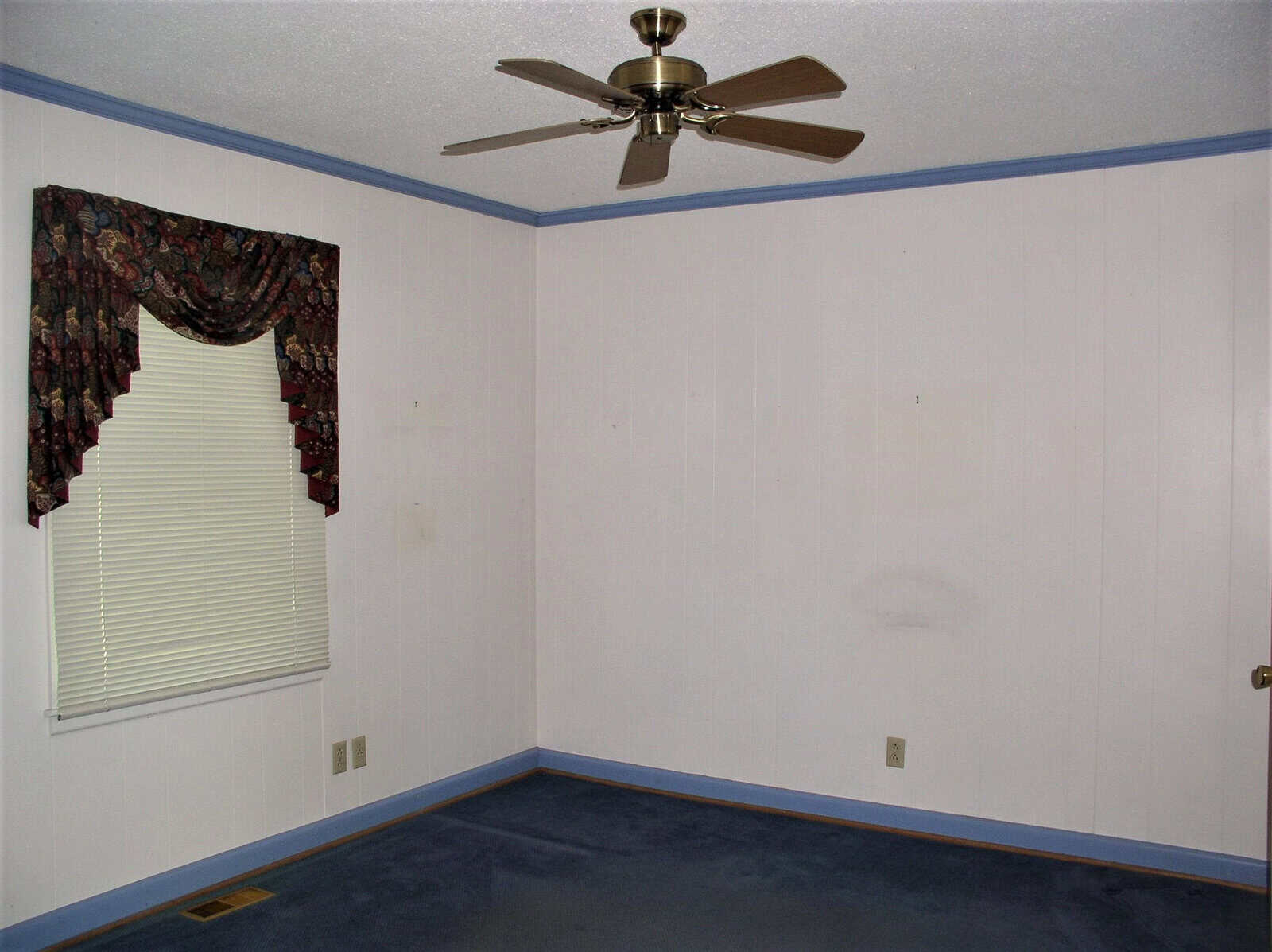 An empty room with white walls, a window with blinds and a patterned valance, a blue carpet, and a ceiling fan.