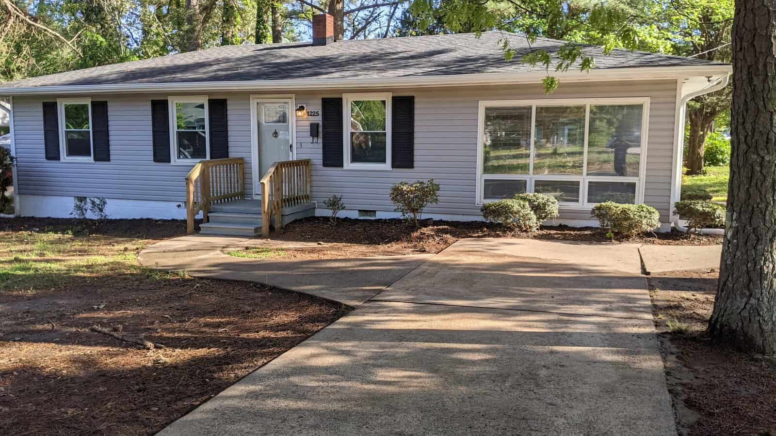 A single-story house with white siding and dark shutters, featuring a small front porch with wooden stairs, surrounded by a freshly mulched garden bed, and a concrete walkway leading to the front door.