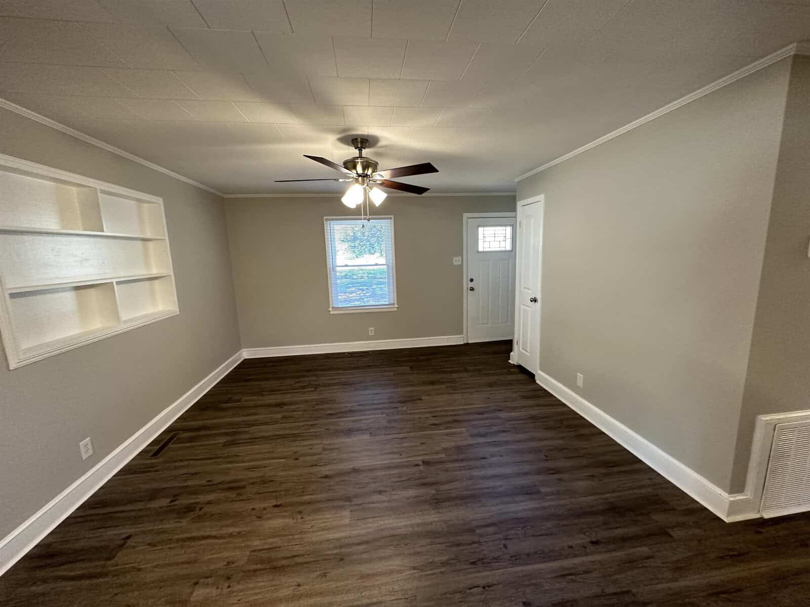 An empty room featuring dark wood flooring, white walls with one accent wall containing built-in shelving, a ceiling fan, and a closed door with a window to the outside.