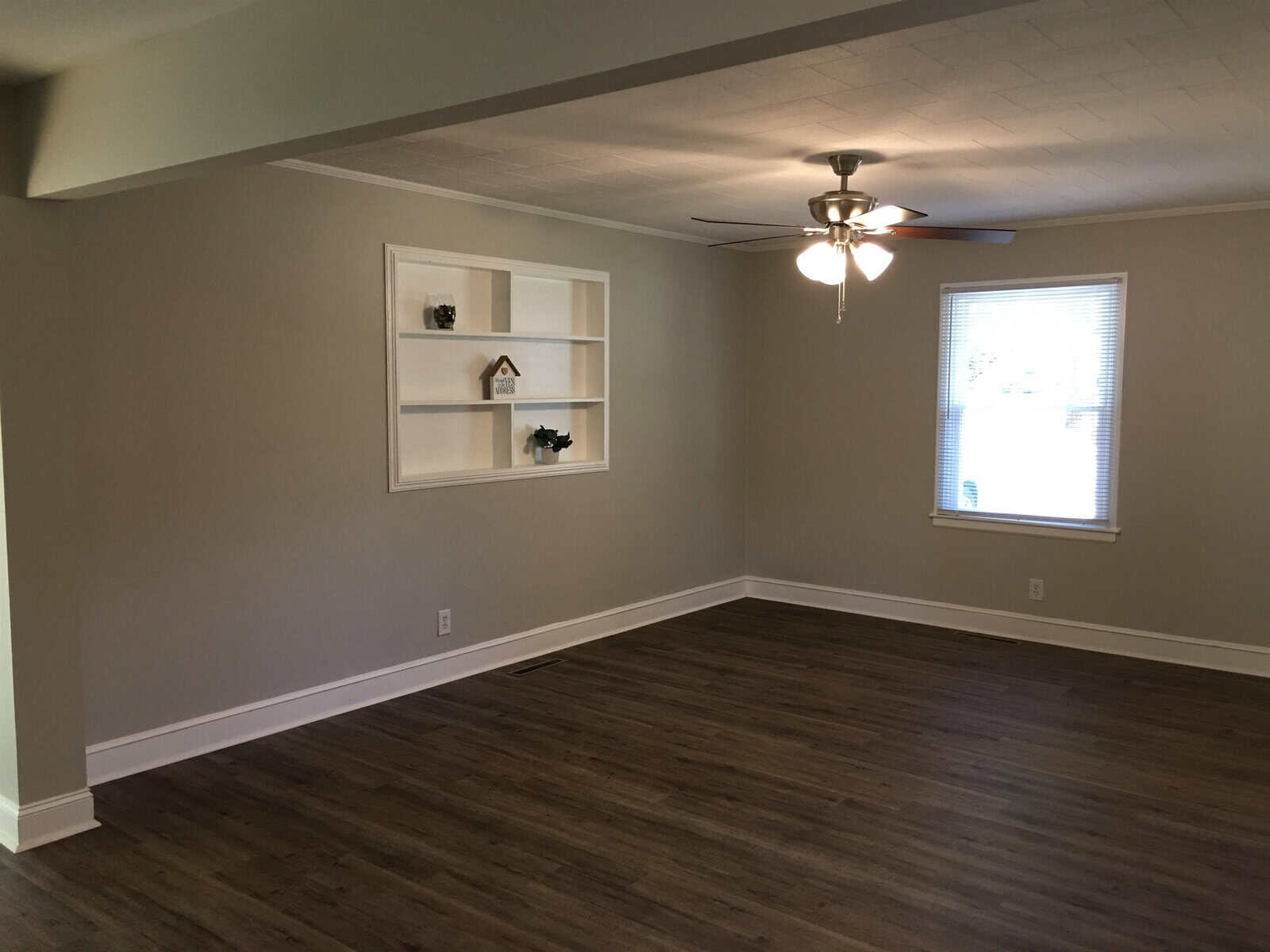 An empty room with beige walls, dark laminate flooring, a ceiling fan with lights, and a built-in white shelving unit with a few decorative items on the shelves. a window provides natural light to the space.
