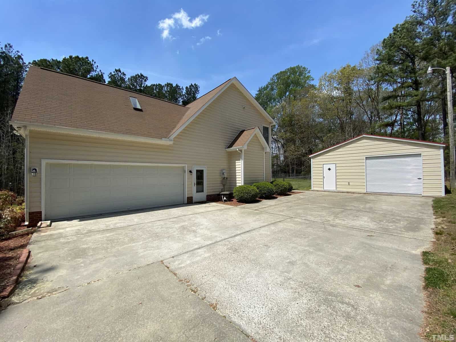 A two-story house with beige siding and a two-car garage, featuring an additional single-car detached garage and a concrete driveway, set against a backdrop of trees and a clear sky.