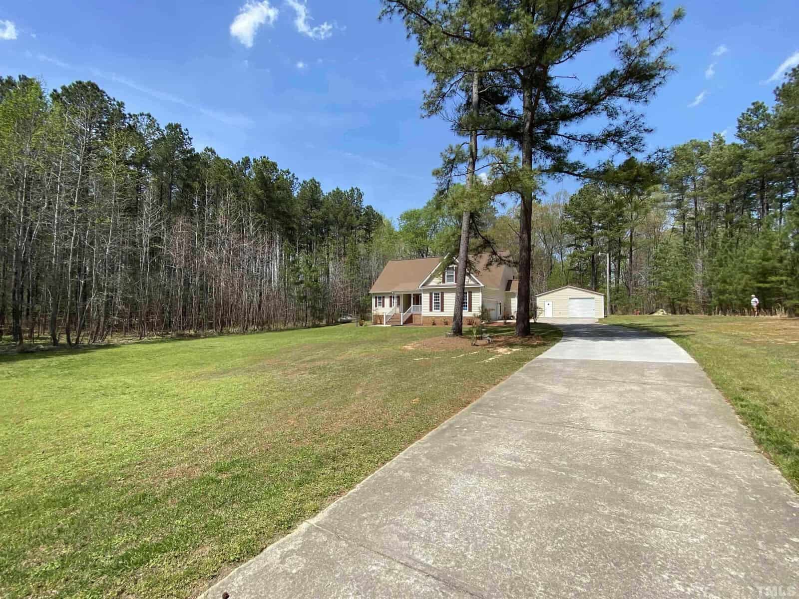 A single-family house with a beige exterior and white trim, featuring an attached garage and a detached storage shed, is set back from a long concrete driveway, surrounded by a well-manicured lawn and a backdrop of tall pine trees under a clear blue sky.