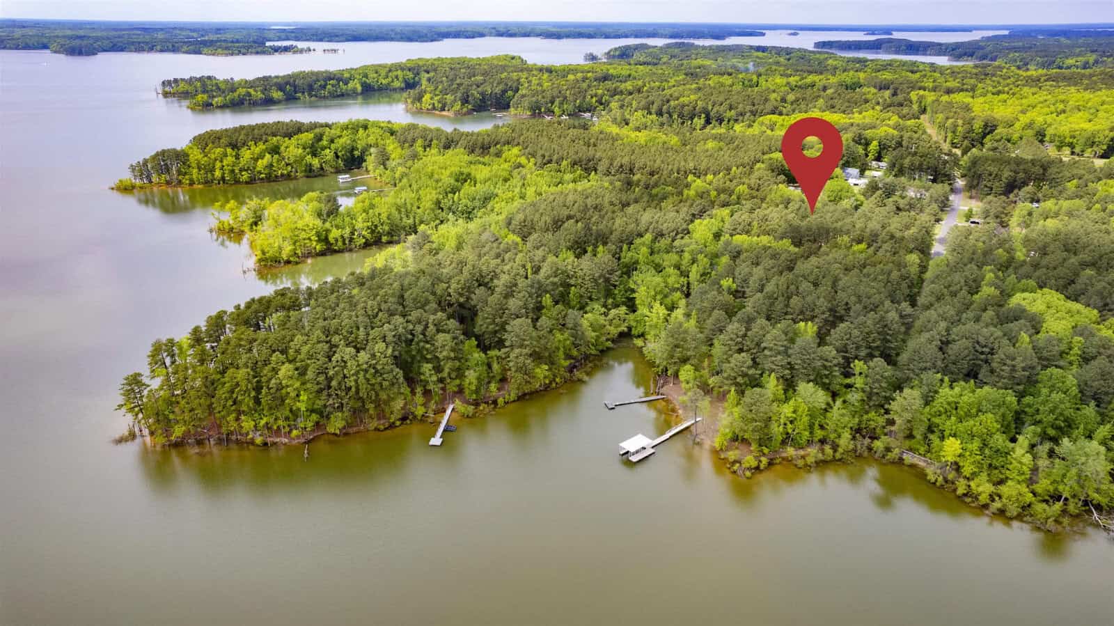 Aerial view of a lush, green, wooded peninsula surrounded by a lake with several docks. A red location pin marks a specific spot among the trees, indicating a point of interest.