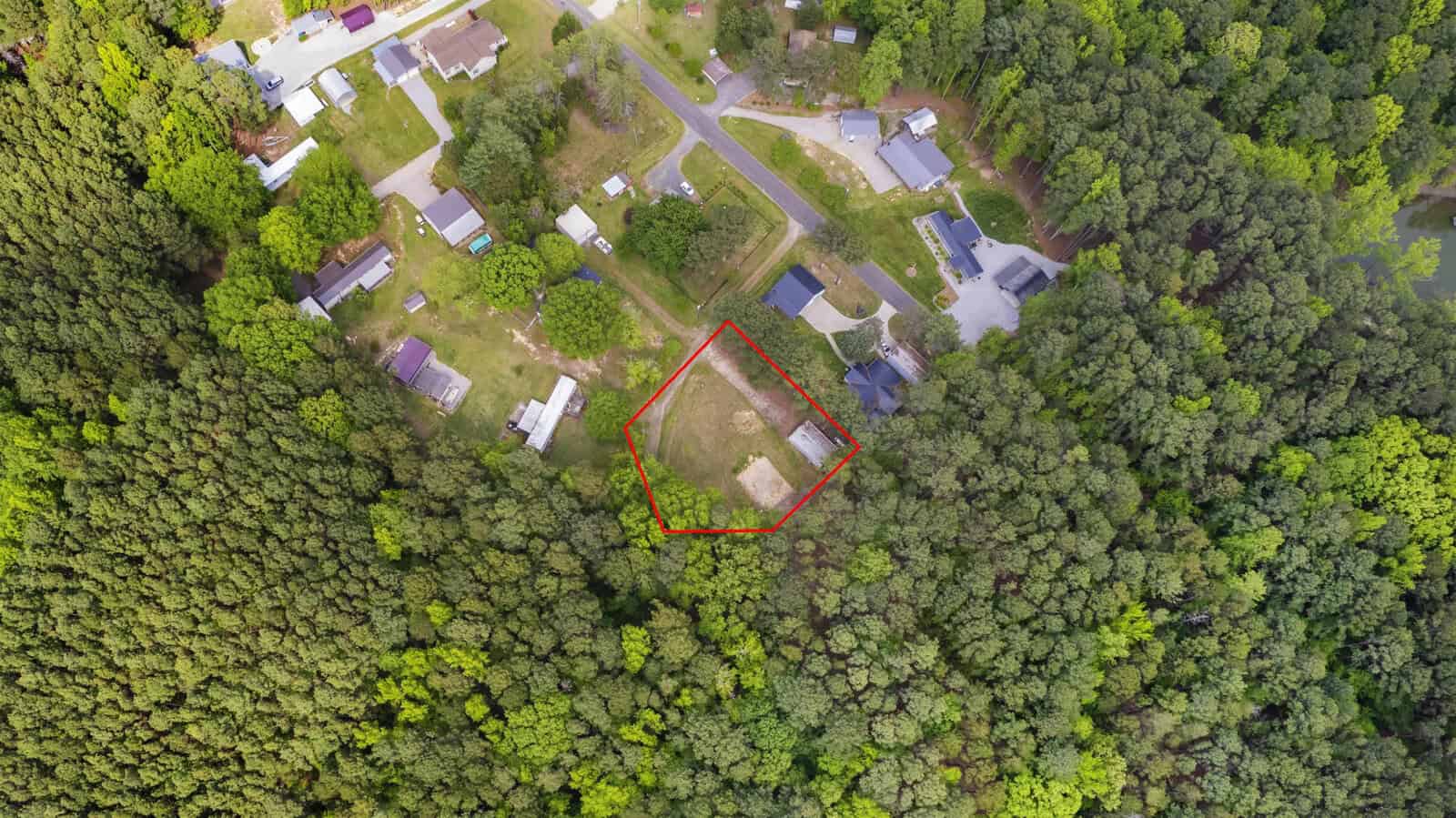 Aerial view of a wooded area with a cleared section outlined in red, surrounded by dense trees and nearby houses along curved roads.