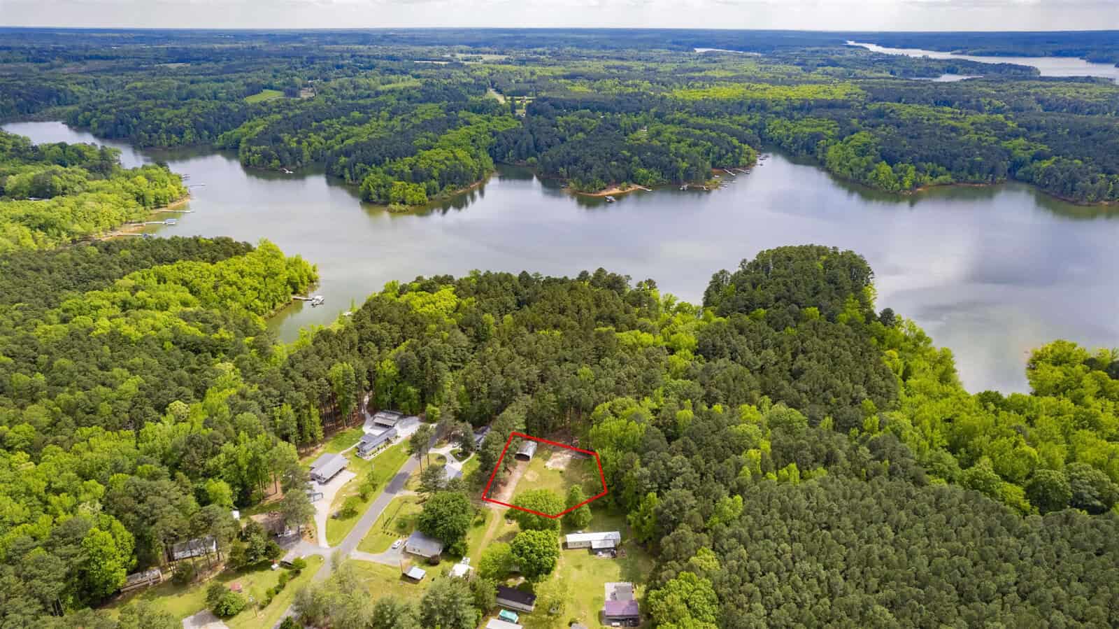Aerial view of a forested lakeside area with scattered houses along a road; one property is outlined in red near the shoreline, surrounded by trees and water.