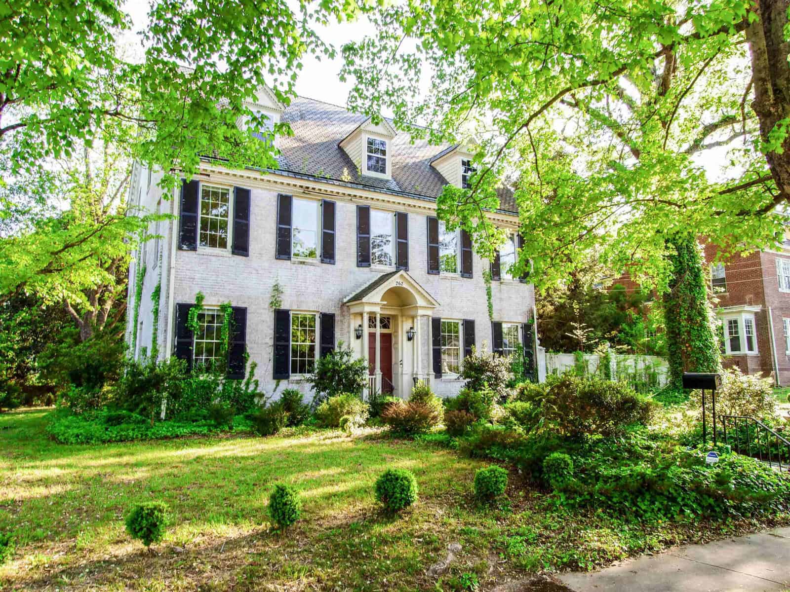 A large two-story white brick house with black shutters, a red door, and surrounded by lush green trees and shrubbery.