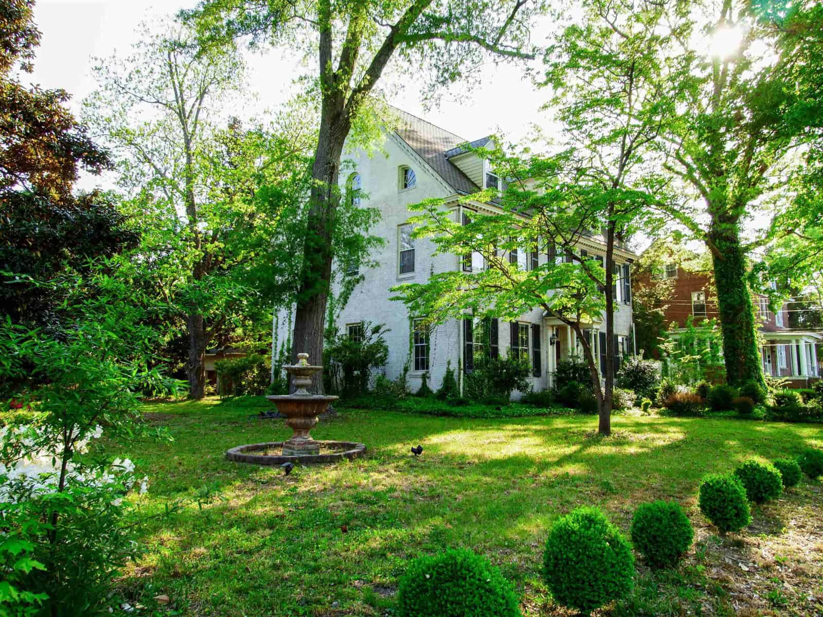 A sunny day casts light on a green, lush garden in front of a large, traditional white house, with a fountain as the centerpiece of the yard.