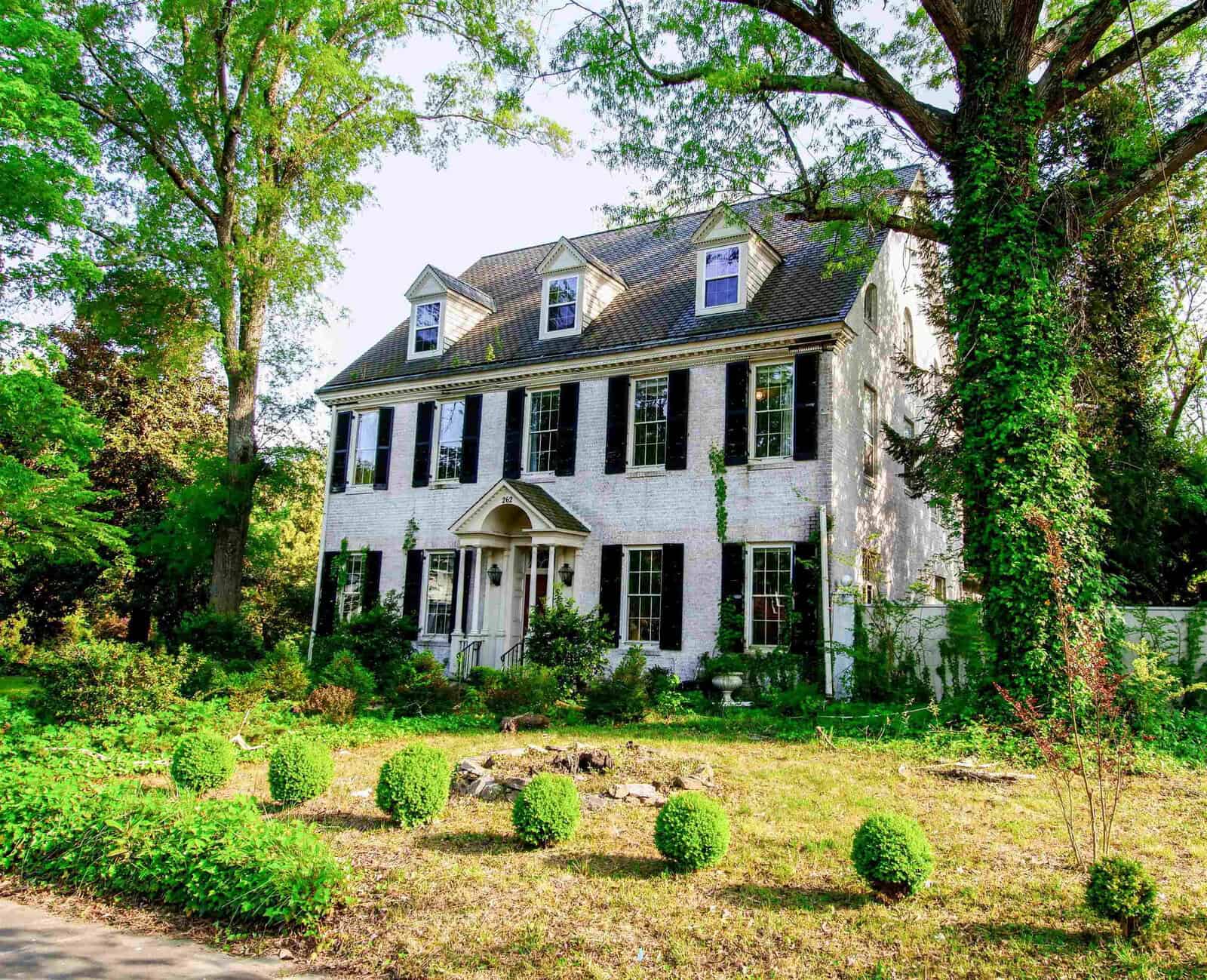 A large two-story white colonial house with a shingled roof, surrounded by greenery and trees, under a clear blue sky.