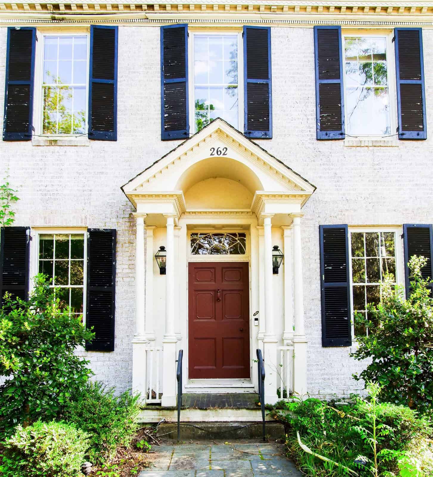 A white brick building featuring a red door with a semicircular transom window, flanked by black shutters and surrounded by greenery.