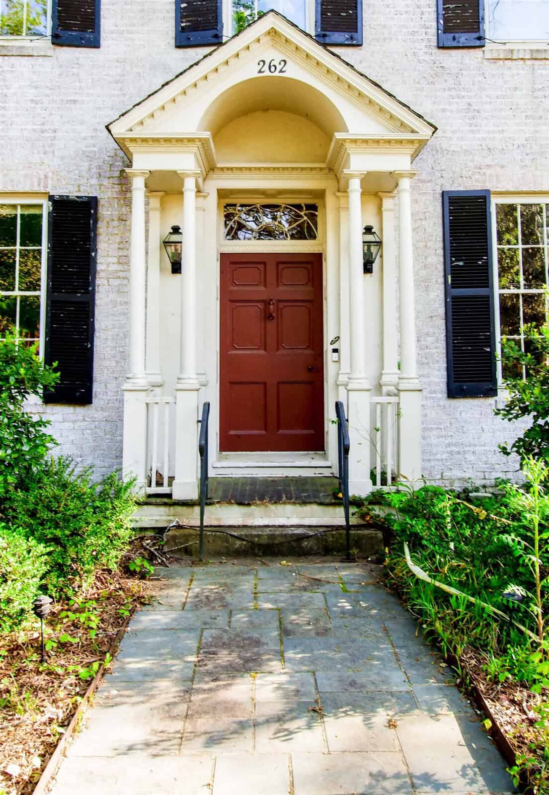 A traditional house entryway featuring a red door, white exterior walls with black shutters, a columned porch, and overgrown greenery flanking a stone pathway.