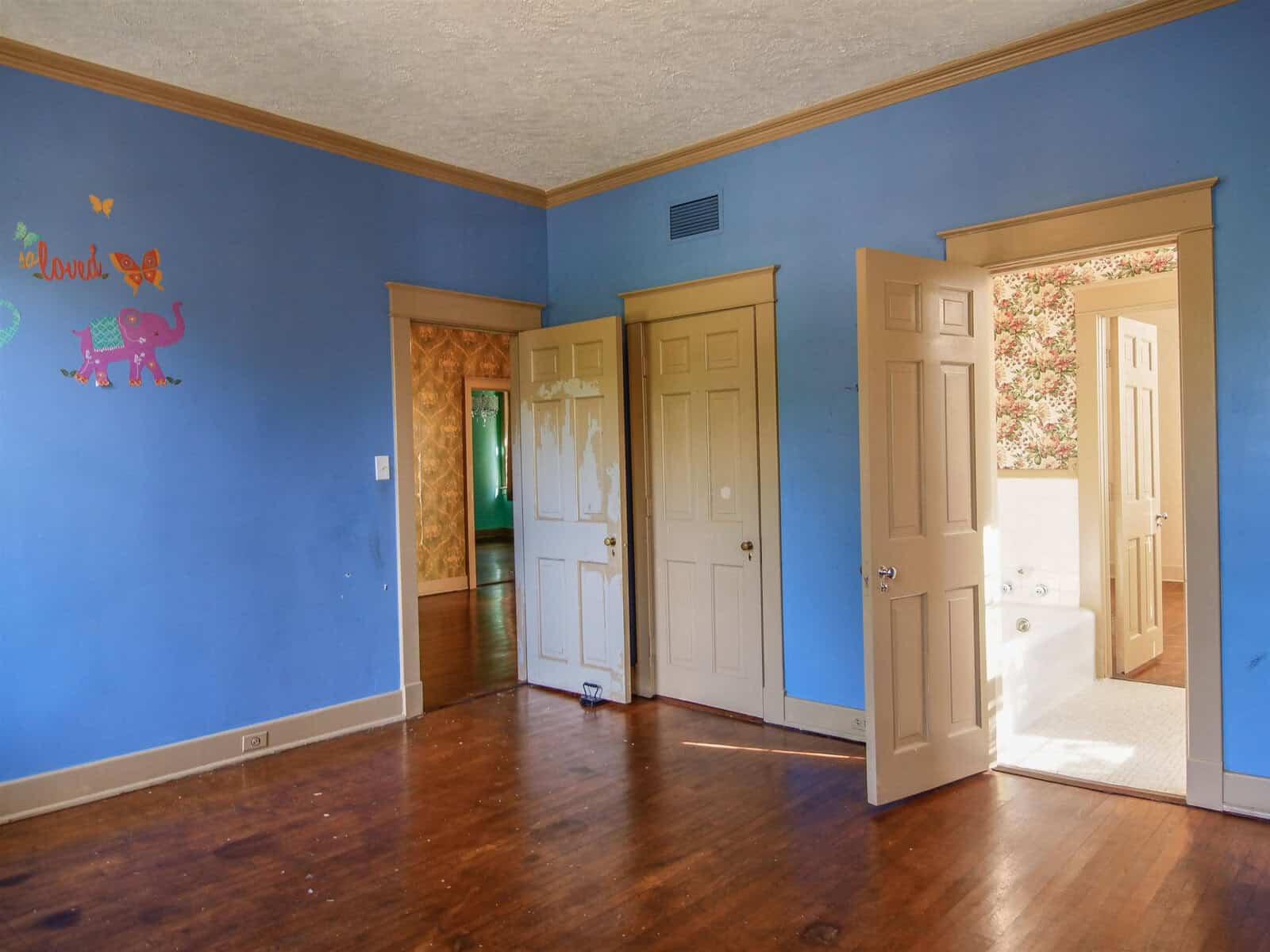 Blue-painted room with wooden floors, children's wall decals, two doors open to adjacent rooms, one revealing a bathroom.