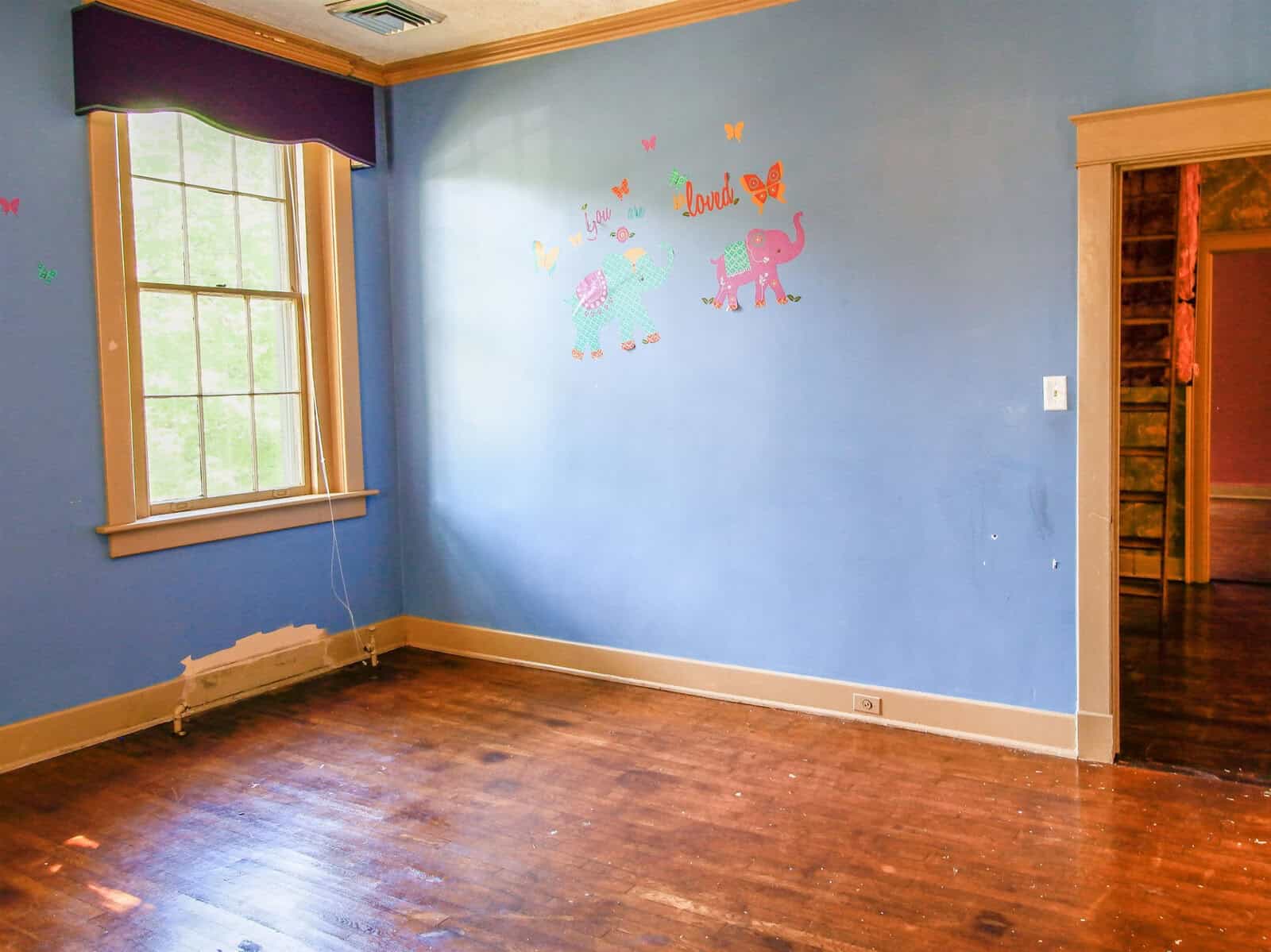 An empty room with hardwood floors, pale blue walls adorned with colorful animal decals, and one window with a view of greenery outside.