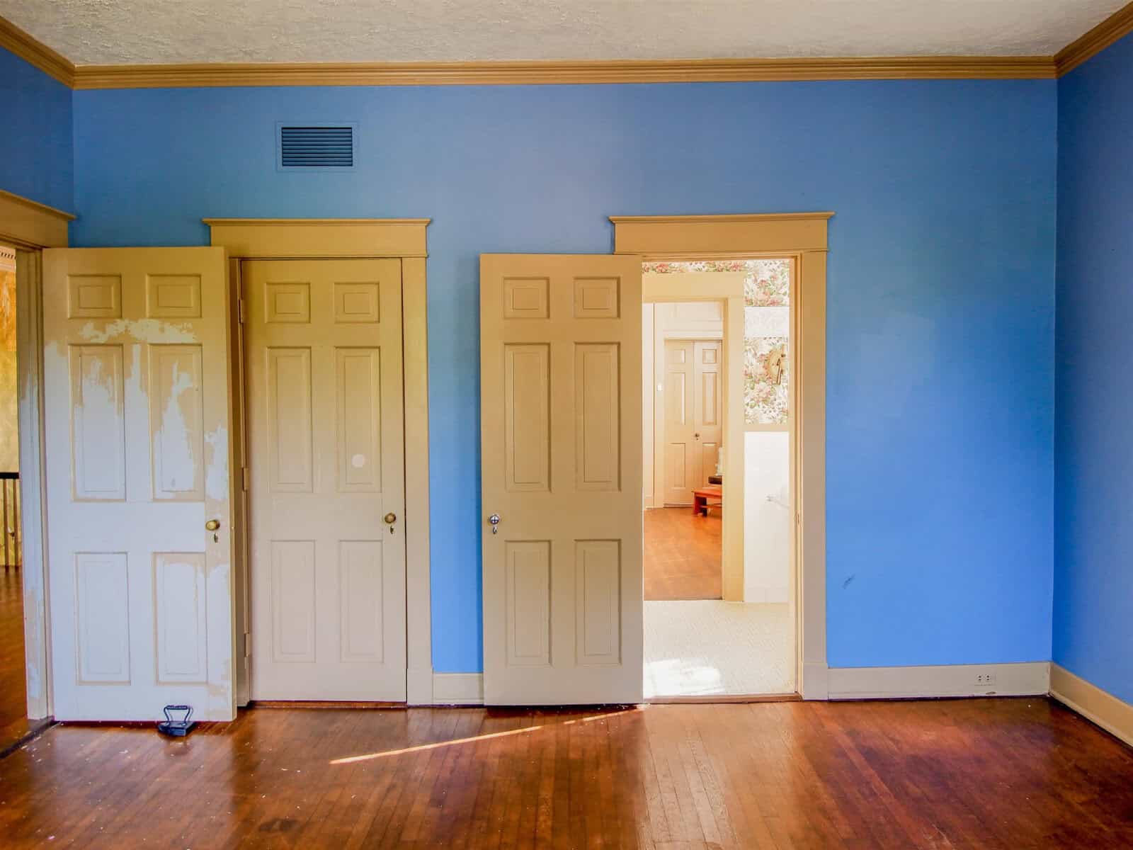 An empty room with blue walls and hardwood floor showing signs of wear and peeling paint on two closed doors, with an open doorway leading to another room.