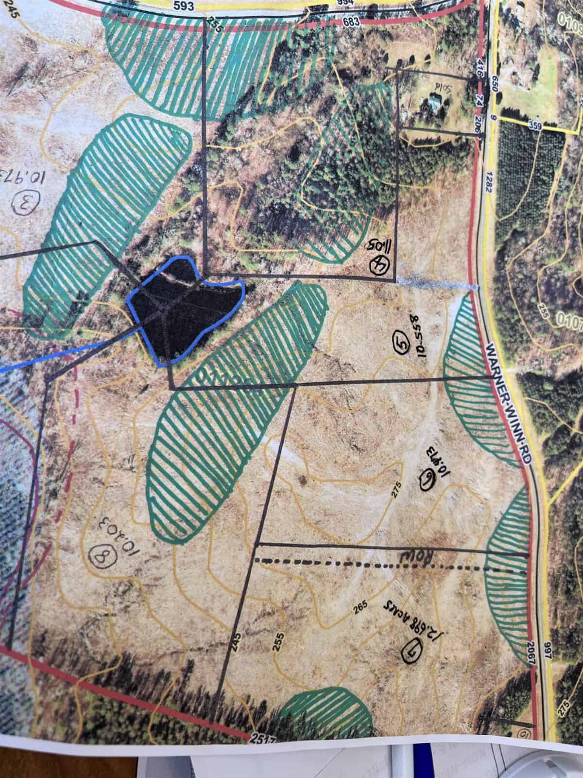 A topographical map displaying various plots of land with green cross-hatched areas, elevation markers, and a blue outline around a water body. Roads and property boundaries are highlighted in black and red. Some handwritten notes are visible around the edges.