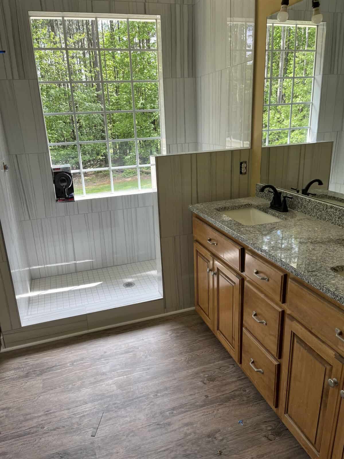 A modern bathroom with wooden cabinets, a granite countertop with dual sinks, large mirrors, and a walk-in shower, featuring tiled walls and a glass door. light streams in from a window overlooking greenery.