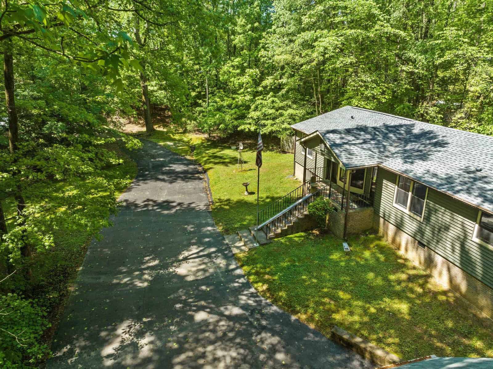 Aerial view of a single-story house with a gray roof, surrounded by lush green trees, featuring a winding driveway and a well-maintained lawn.