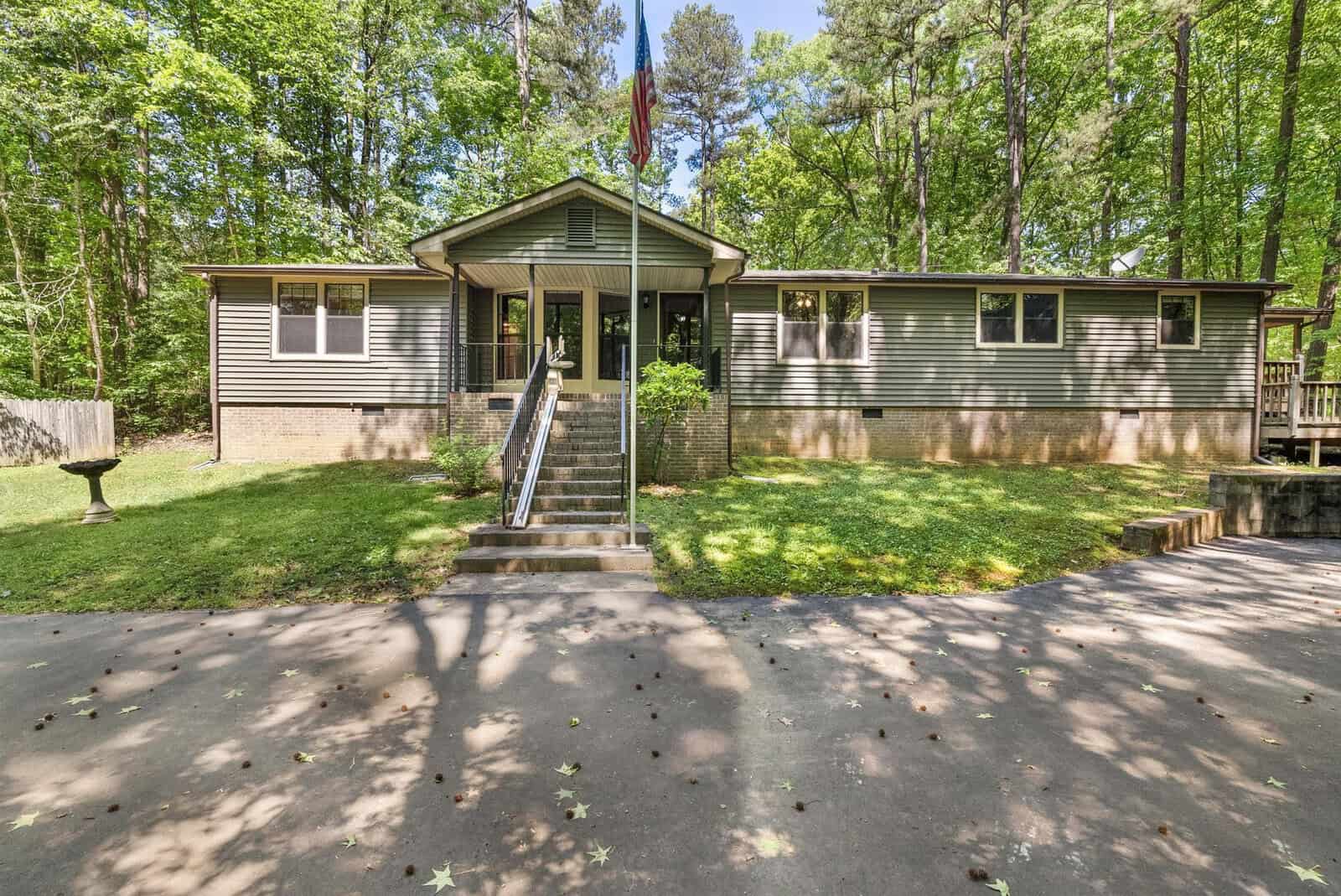 This is an image of a single-story house with a brown exterior and a gabled roof, featuring a front porch with stairs and a railing. the house is surrounded by a lawn with scattered leaves and trees, and there is a concrete driveway in the foreground.