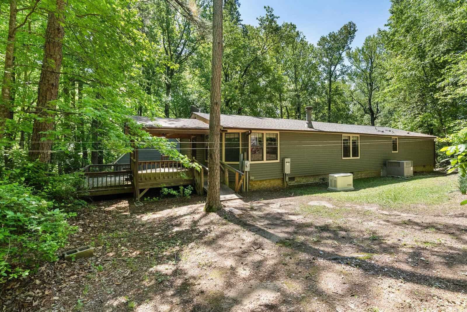A single-story house with beige siding is nestled among trees, featuring a small deck and a prominent air conditioning unit on the side.