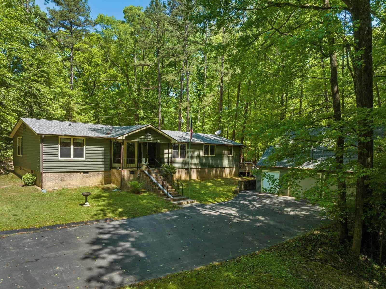 A single-story residential home with a dark roof and light-colored siding, featuring an attached garage, is surrounded by tall, lush green trees. a paved driveway leads to the garage and a set of stairs leads to the main entrance.