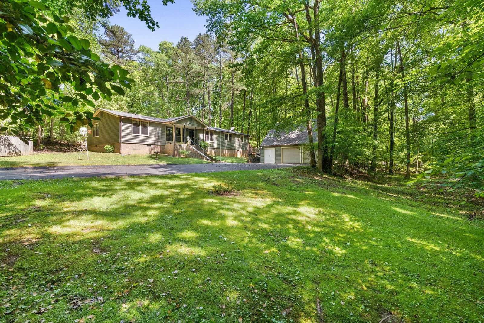 A single-story house with an attached garage set back in a lush, wooded area with a driveway leading to a detached secondary garage.