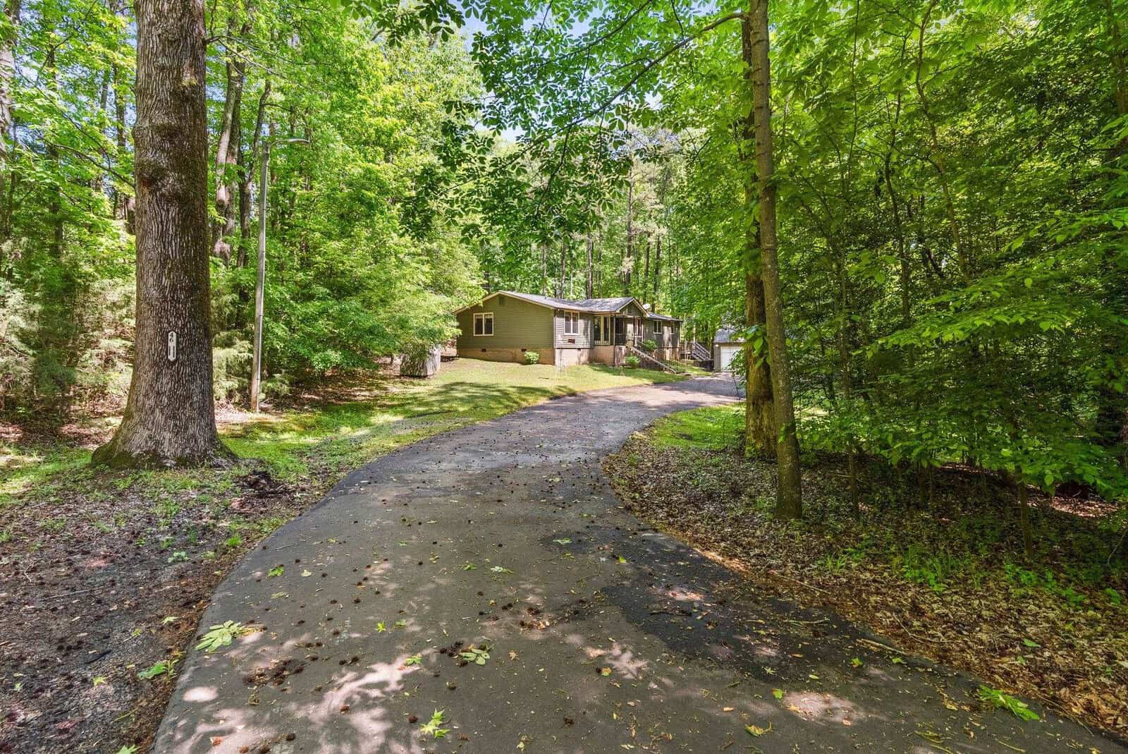A single-story house with a green exterior set back from a curved driveway, surrounded by lush trees in a forested area.