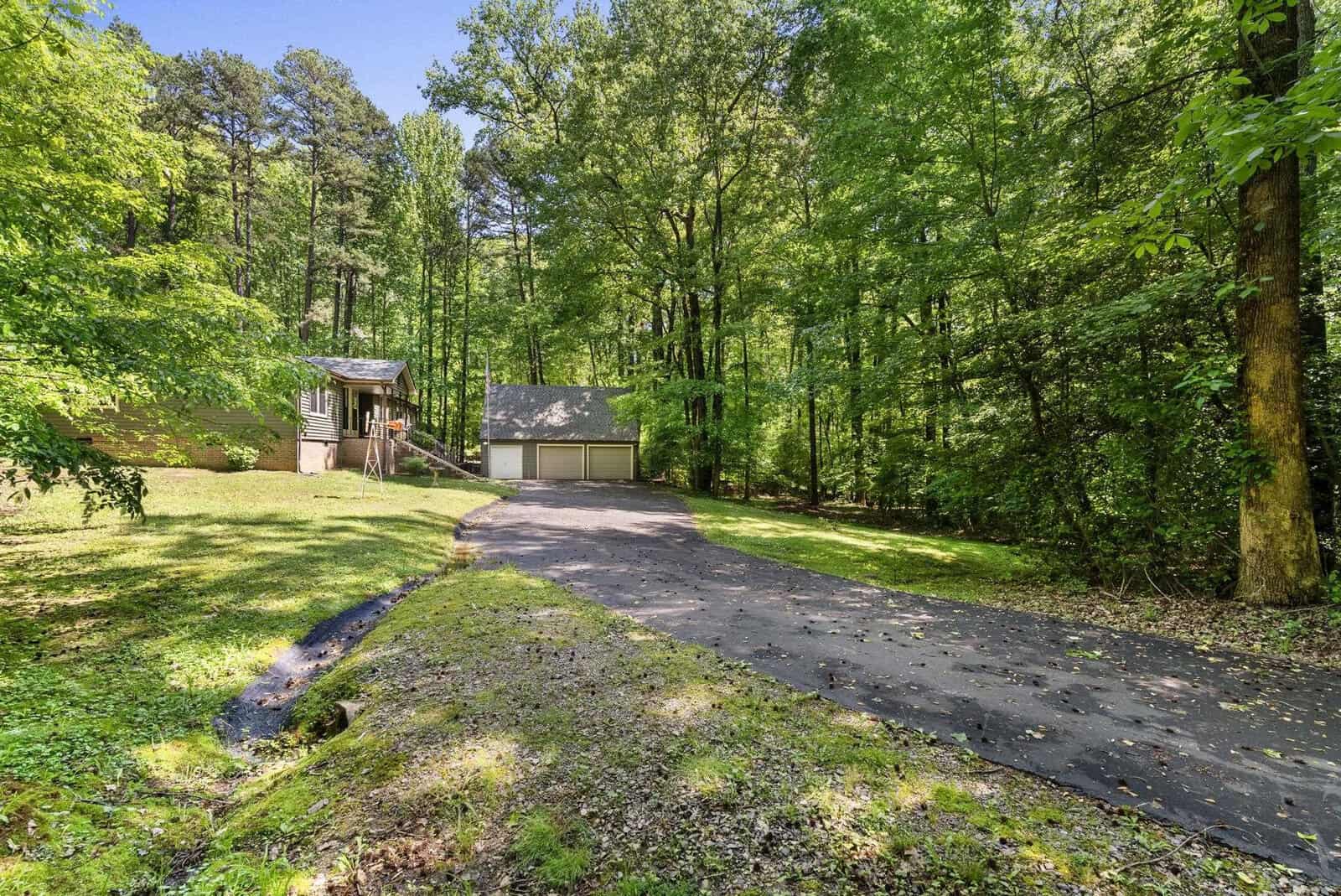A residential property featuring a driveway leading to a detached garage, surrounded by a lush green woodland setting. the house is partially visible to the left with an elevated deck.
