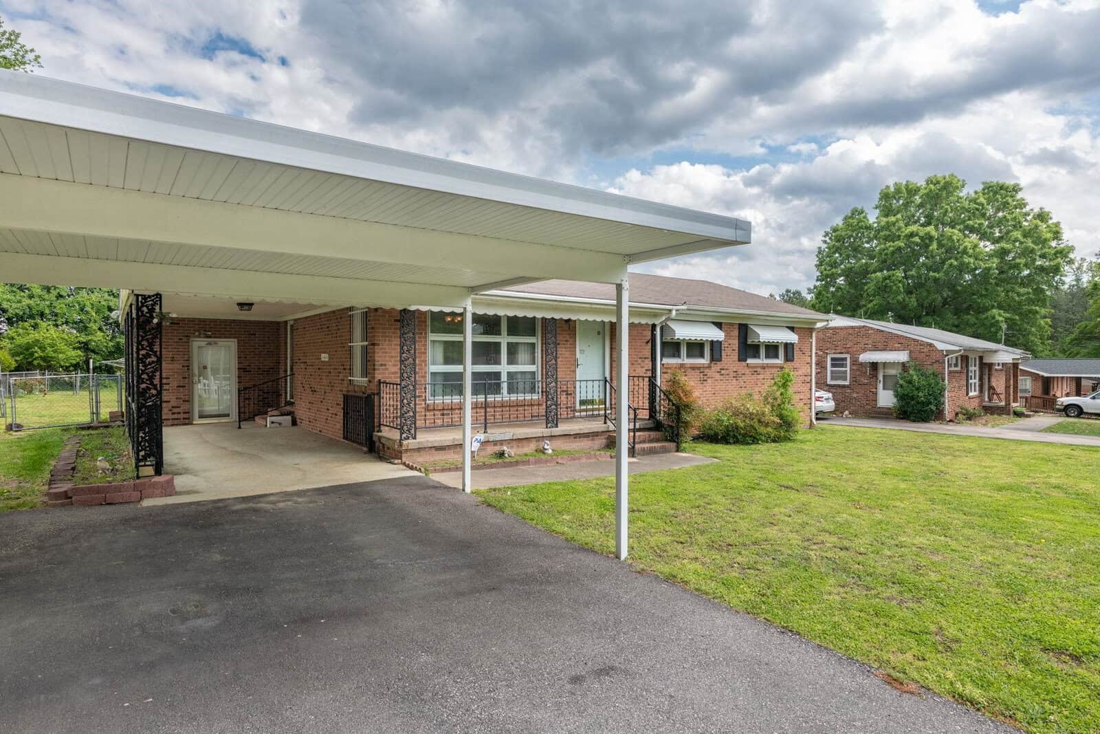A single-story brick residential building with an attached carport and a small covered porch area, surrounded by a lawn with scattered trees under a partly cloudy sky.