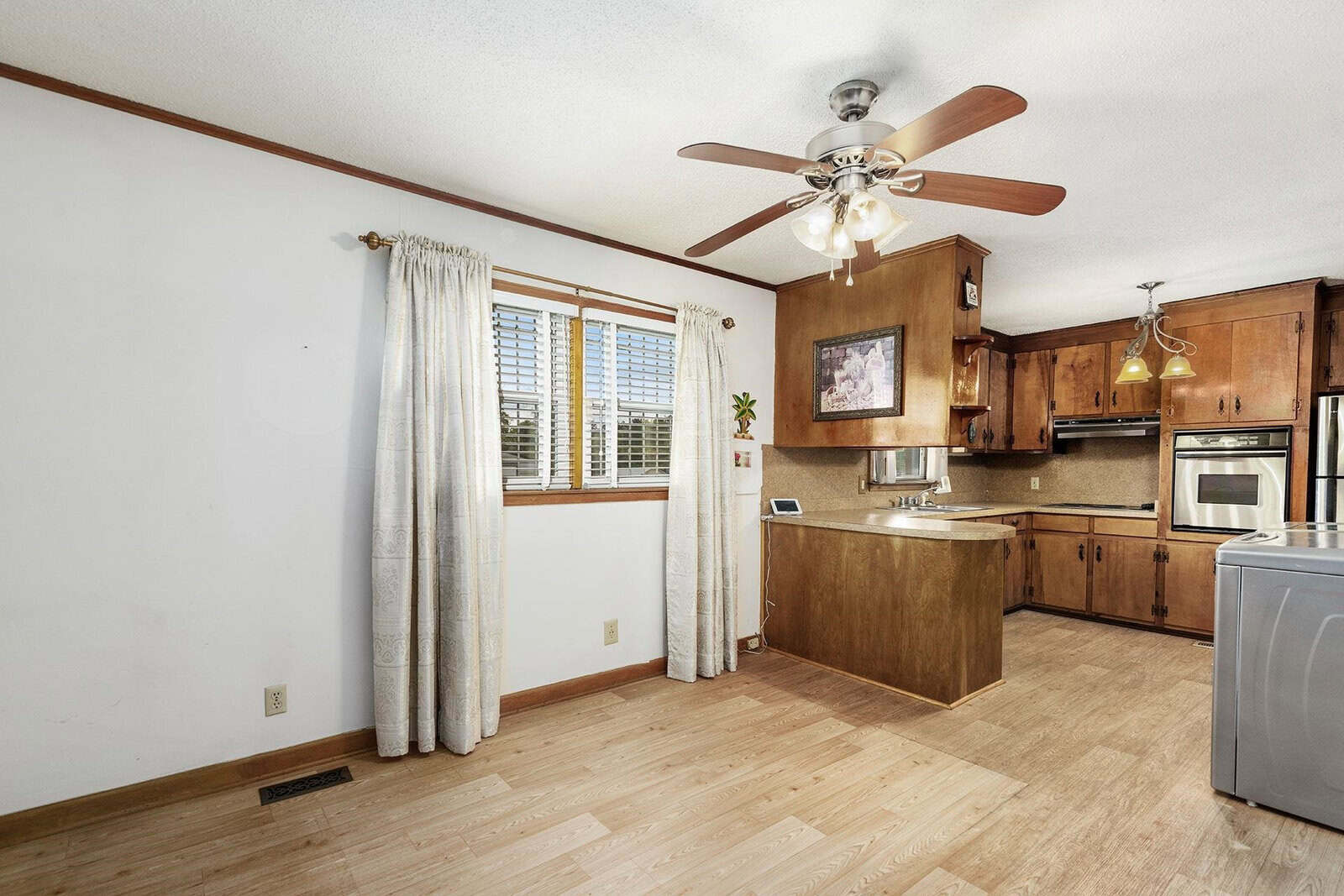 A well-lit, empty kitchen with wooden cabinets, hardwood flooring, a ceiling fan, and a window with white curtains.
