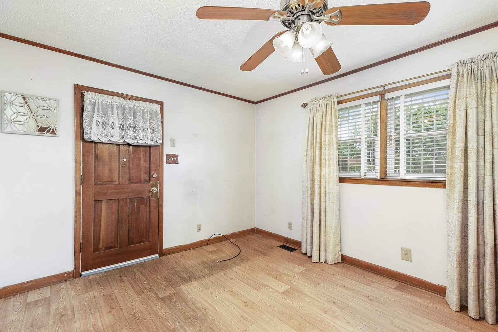 Empty room with laminate flooring, a wooden door, a ceiling fan, and a window with blinds and curtains.