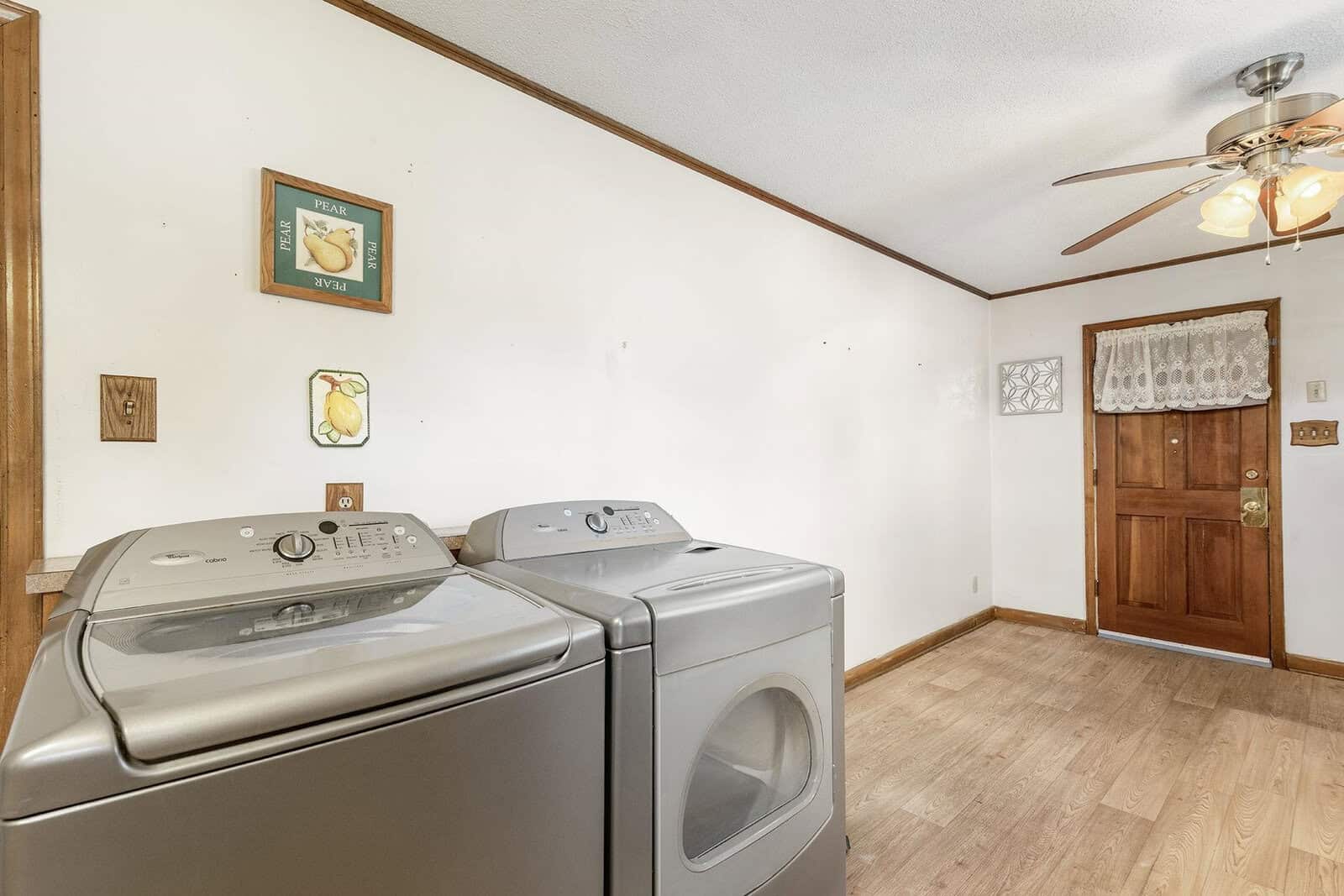 A laundry room with a washer and dryer in the foreground, a framed picture on the wall, and a wooden door at the back. a ceiling fan with lights is overhead.