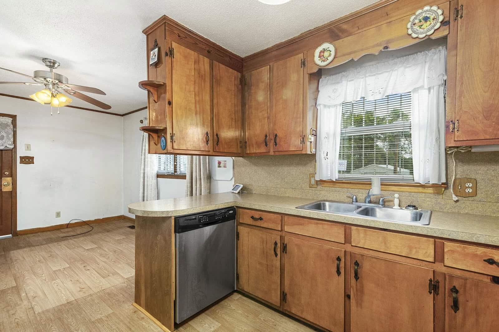 A wooden-finished kitchen interior featuring a double basin sink below a window, cabinets, a dishwasher, and a fan overhead, with tiled countertops and laminate flooring.
