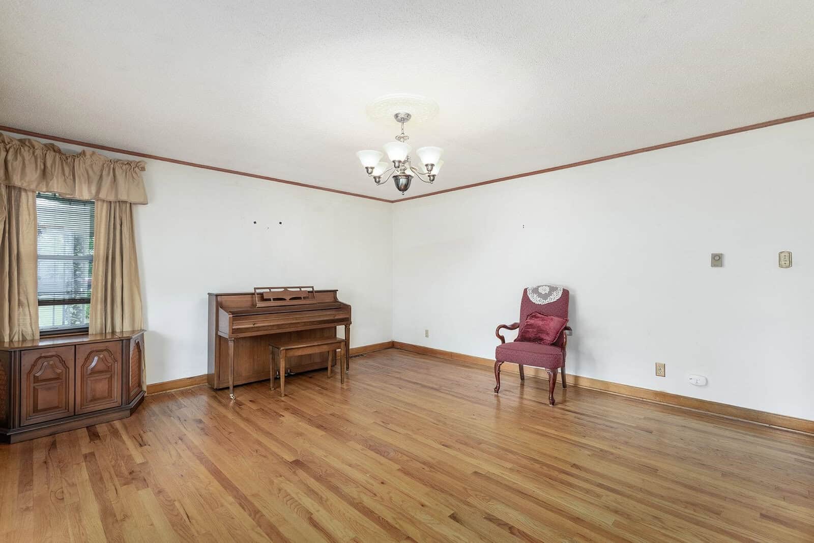 A sparsely furnished room with hardwood flooring, featuring a wooden piano, a vintage armchair, and a side cabinet under a window with draped curtains.