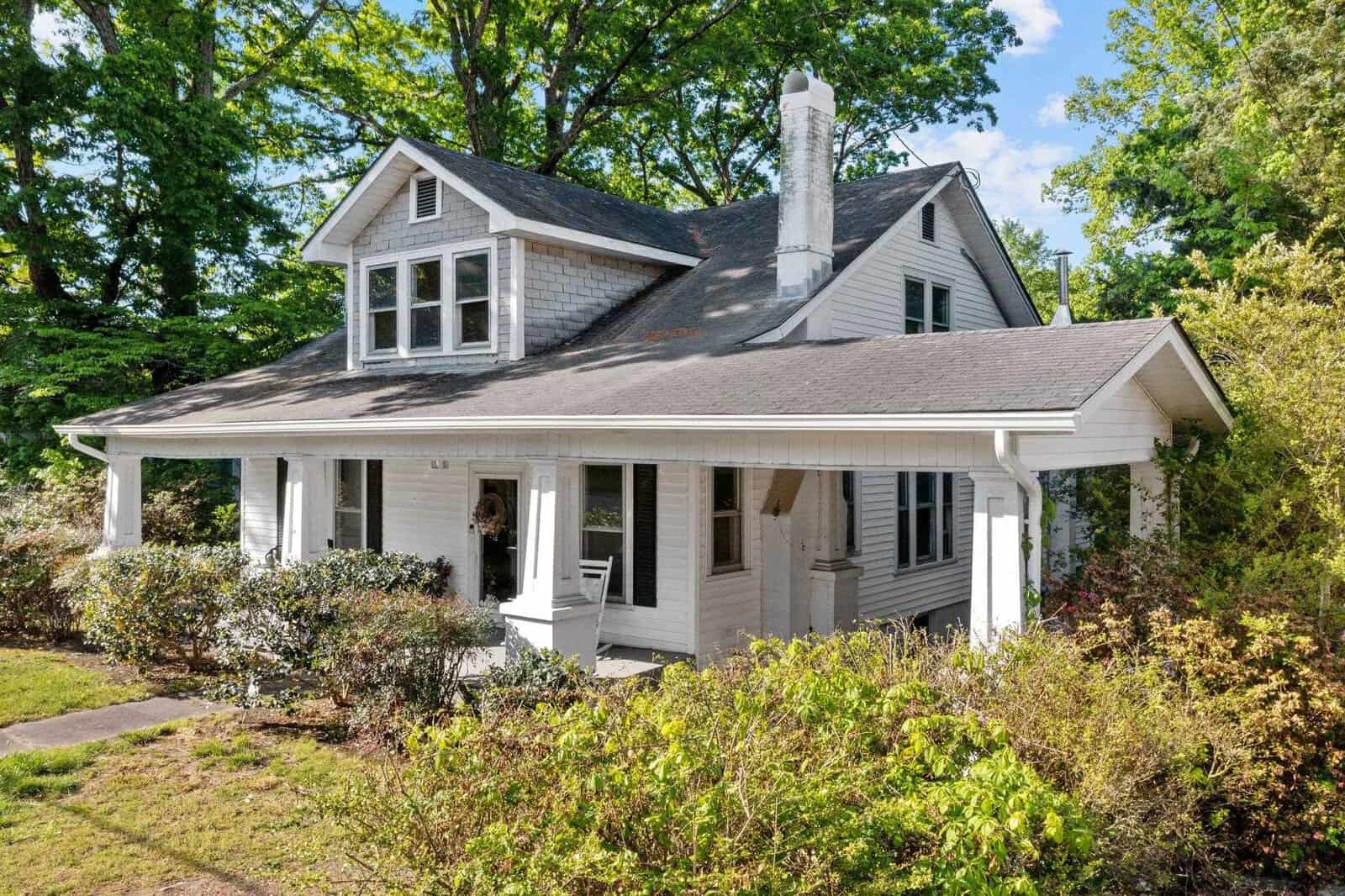 A quaint one-and-a-half-story house with a covered porch, surrounded by greenery on a sunny day.
