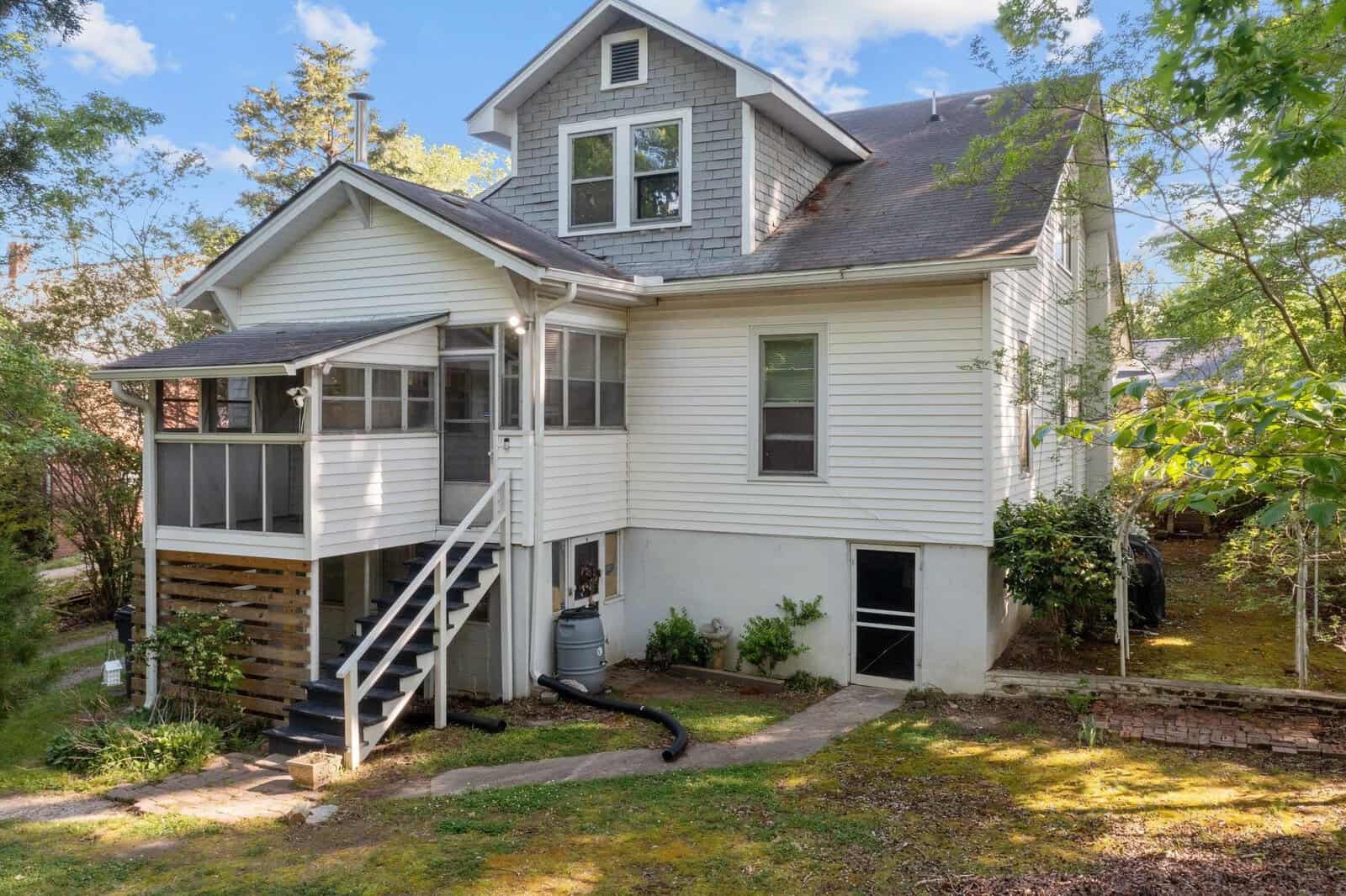 A two-story white house with a screened porch, external staircase, and adjacent greenery on a sunny day.