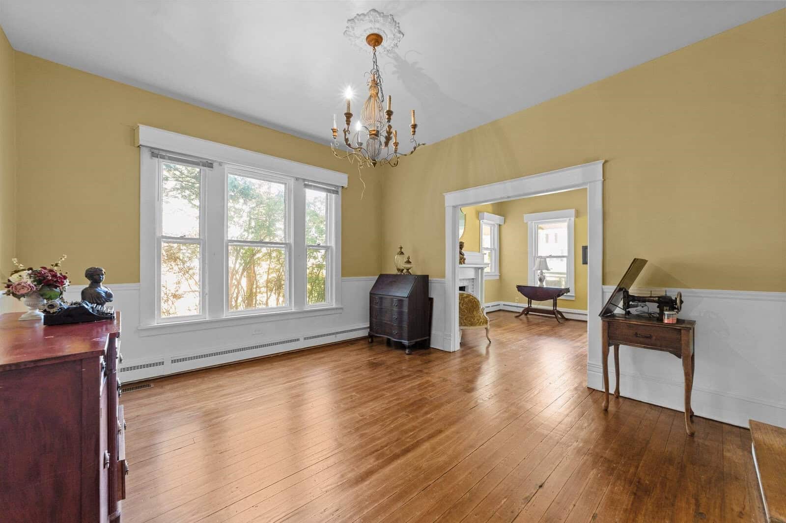 A spacious, well-lit room with yellow walls, hardwood floors, and large windows. a chandelier hangs from the ceiling and a few pieces of furniture are arranged around the perimeter of the room.
