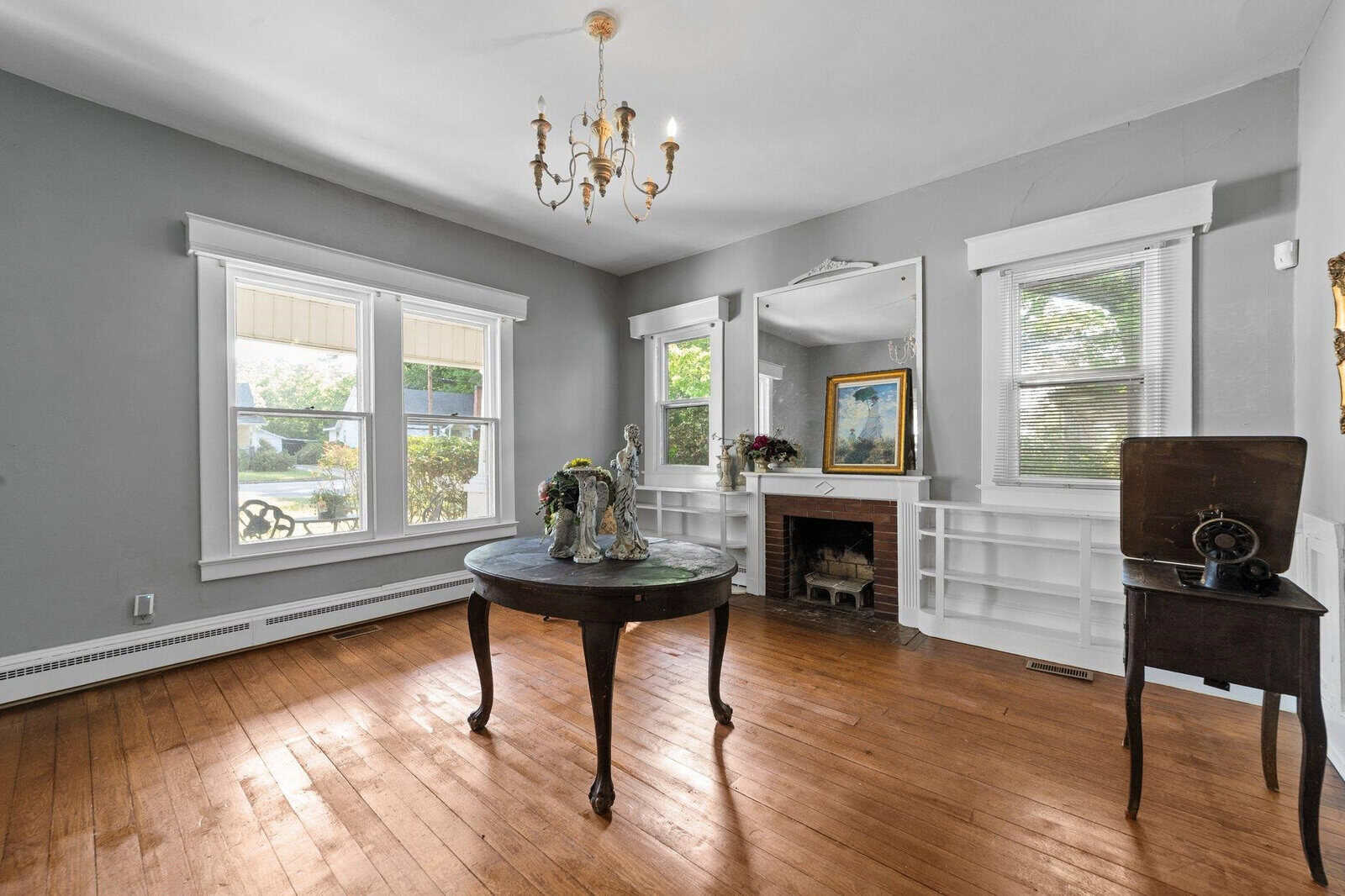 An empty living room with hardwood floors, light gray walls, and white trim. the room features a round wooden table in the center, a fireplace with a white mantle, built-in shelving, two windows providing natural light, and a chandelier. there is an antique-looking phonograph on a small table near one of the windows.