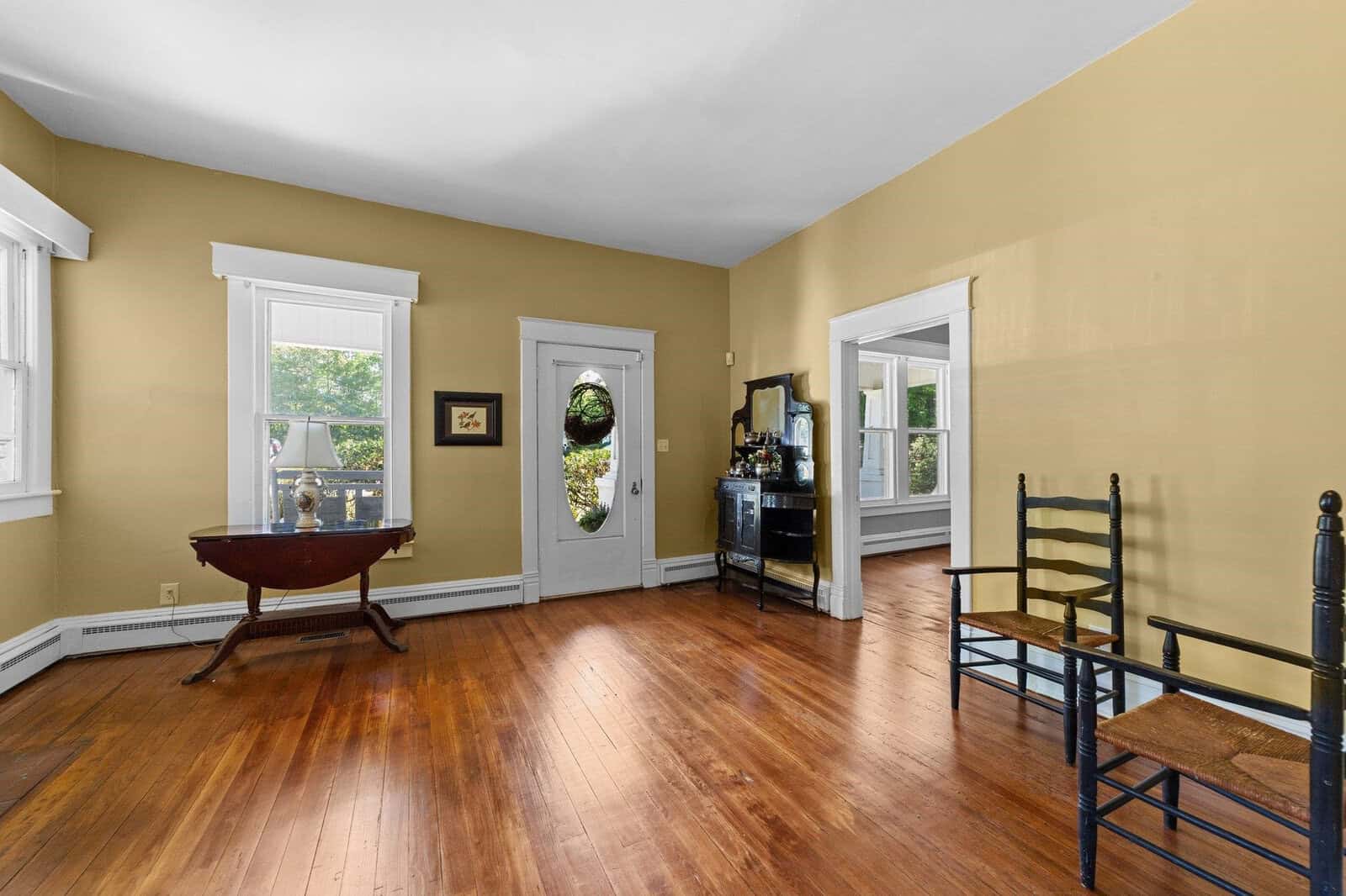 Empty living room with hardwood floors, pale yellow walls, two windows, a glass-paneled door, and minimal furniture including a round table and two chairs.