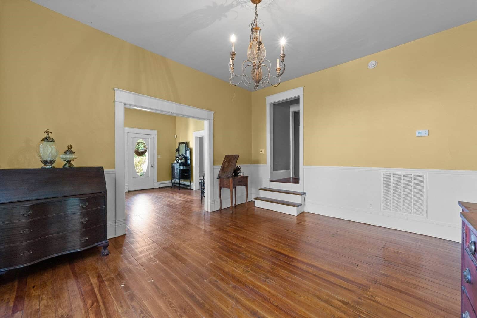 Empty living room with hardwood floors, yellow walls, and a chandelier, featuring a dark wooden chest, a lamp on a small table, and open doorways leading to other rooms.