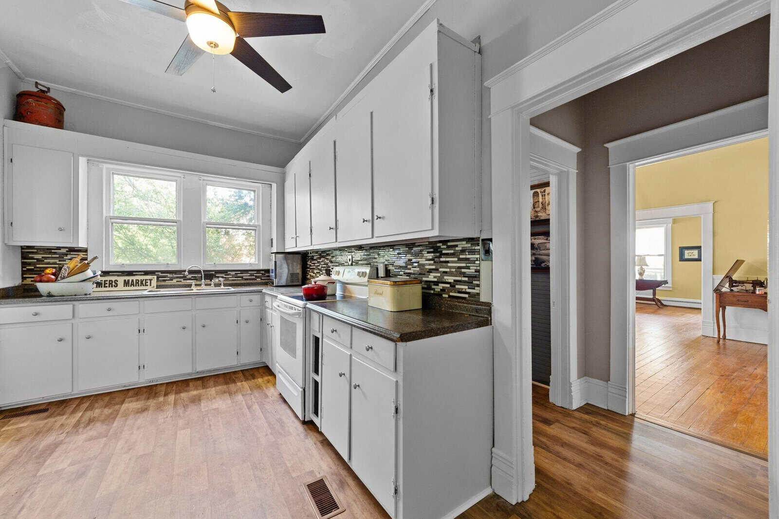 A spacious kitchen featuring white cabinetry, stainless steel appliances, a mosaic backsplash, and hardwood flooring, with a view into adjoining rooms.