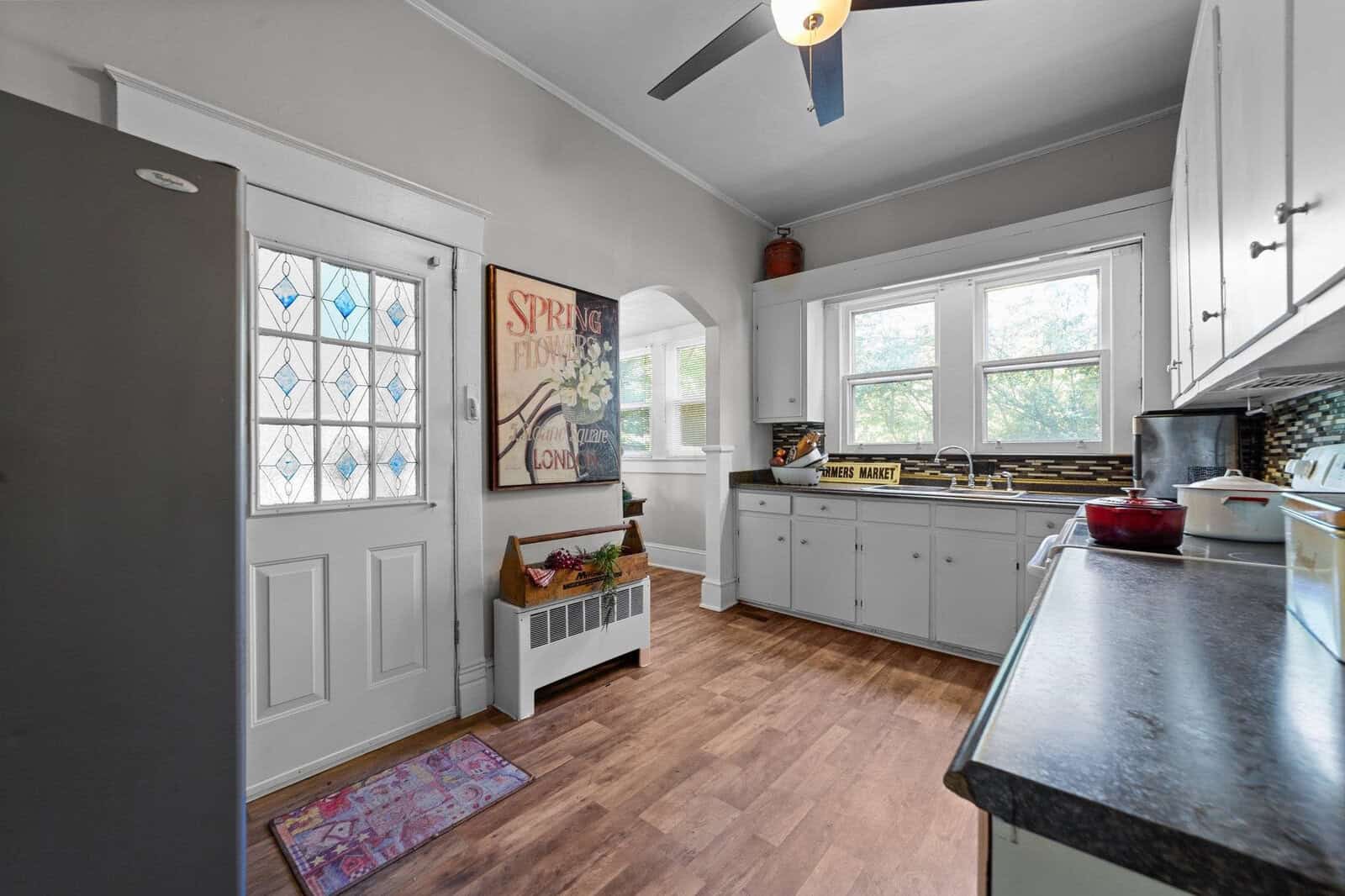 A bright, modern kitchen with white cabinetry, stainless steel appliances, and a hardwood floor. the room includes a ceiling fan, a window above the sink, and decorative touches such as a sign that says "spring" and a basket of flowers on a bench by the door.