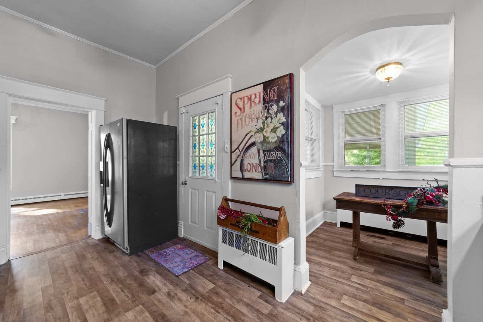 An empty, bright room featuring a hardwood floor, with a black front door on the left and a white bench under a window on the right. decorative elements include a colorful floor mat, large floral artwork on the wall next to the door, and a small table adorned with a floral arrangement and a sign that reads "courage.